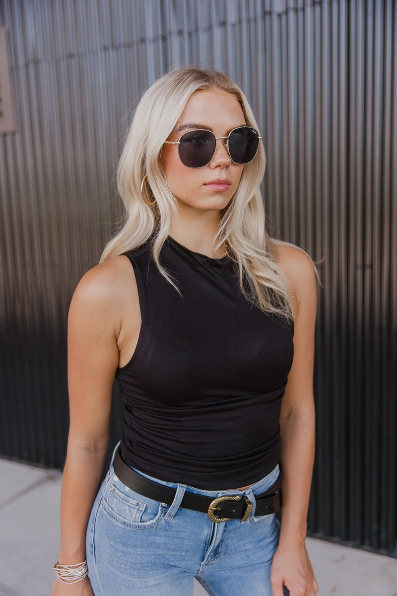 Front view of model wearing the True Story Mock Neck Tank in Black that has black knit fabric, a high neckline, and a sleeveless design. Top is tucked into jeans.