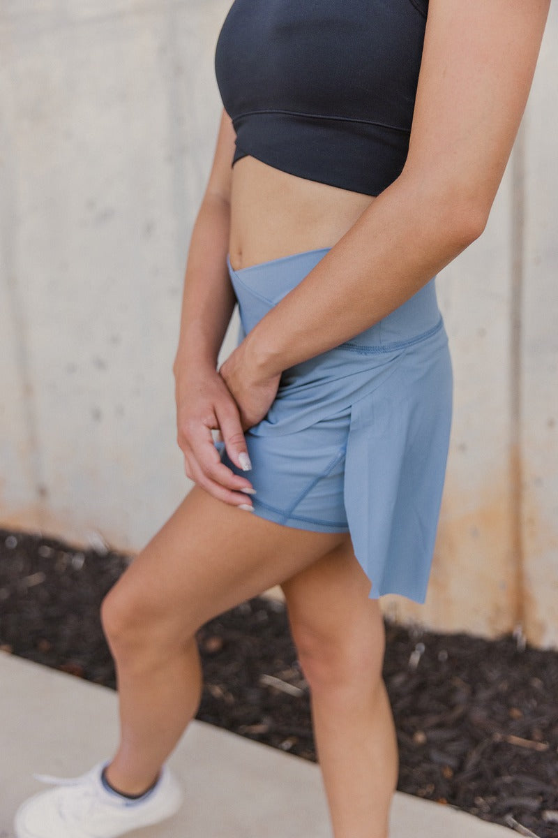 Side view of model wearing the Step It Up Skort in Denim which features dusty blue fabric, shorts lining, a v overlap waistline, slits on each side, and a mini-length hem.