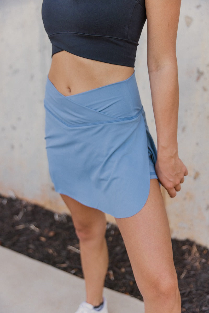 Frontal side view of model wearing the Step It Up Skort in Denim which features dusty blue fabric, shorts lining, a v overlap waistline, slits on each side, and a mini-length hem.