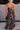 Full body back view of model wearing the Beautiful Mess Floral Dress which features black, orange, fuchsia, teal, aqua, green and white fabric, floral print, midi dress, two-tiered body, square neckline, adjustable straps and monochromatic back zipper wit