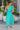 Back view of model wearing the Cabana Bay Midi Dress which features turquoise fabric, midi length, a tie around the waist, turquoise lining, one shoulder ruffle strap, and a monochromatic side zipper with a hook closure.