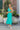 Full body view of model wearing the Cabana Bay Midi Dress which features turquoise fabric, midi length, a tie around the waist, turquoise lining, one shoulder ruffle strap, and a monochromatic side zipper with a hook closure.