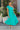 Full body view of model wearing the Cabana Bay Midi Dress which features turquoise fabric, midi length, a tie around the waist, turquoise lining, one shoulder ruffle strap, and a monochromatic side zipper with a hook closure.
