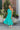 Full body view of model wearing the Cabana Bay Midi Dress which features turquoise fabric, midi length, a tie around the waist, turquoise lining, one shoulder ruffle strap, and a monochromatic side zipper with a hook closure.