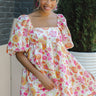 Front view of model wearing the Floral Fields Babydoll Dress that has white sheen fabric with an orange, hot pink, light green, and light pink floral print, a square neck, and 3/4 puff sleeves