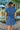 Back view of model wearing the East Side Denim Dress that has dark wash denim fabric, mini length, pockets, a silver button up front, a collared neck, a tie around the waist, and short sleeves.