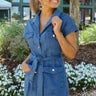 Front view of model wearing the East Side Denim Dress that has dark wash denim fabric, mini length, pockets, a silver button up front, a collared neck, a tie around the waist, and short sleeves.