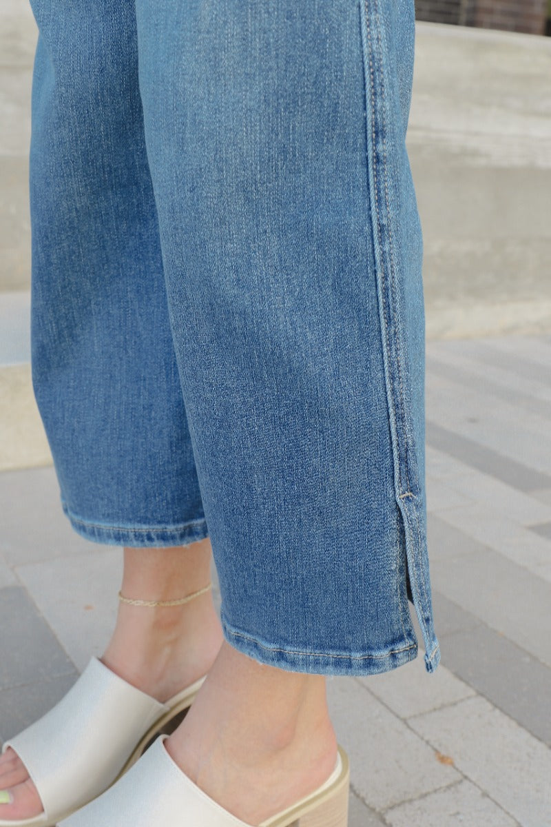 Close up view of model wearing the Rooted Denim: Breaking Rules Cropped Flare which features medium denim wash fabric, cropped flare leg, front zipper with button closure, two front pockets, two back pockets, belt loops and distressed details.