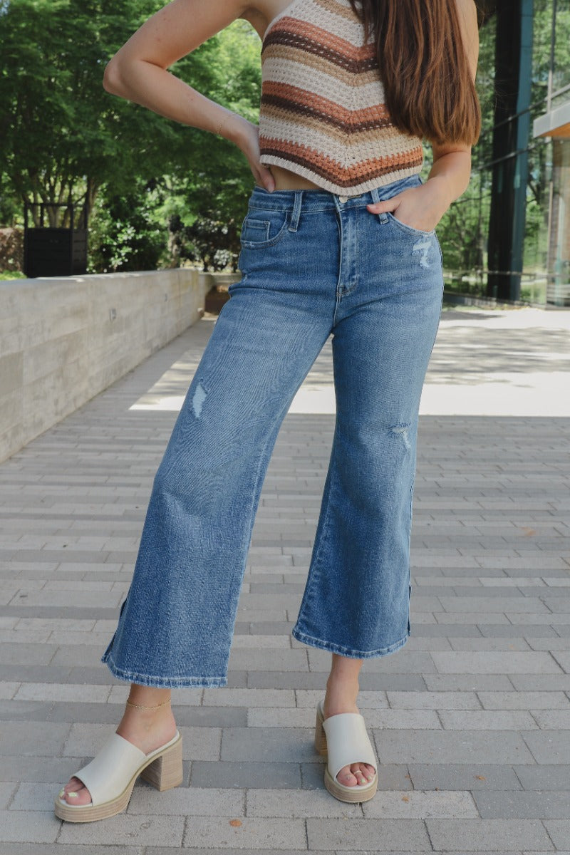Front view of model wearing the Rooted Denim: Breaking Rules Cropped Flare which features medium denim wash fabric, cropped flare leg, front zipper with button closure, two front pockets, two back pockets, belt loops and distressed details.
