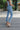 Side view of model wearing the Rooted Denim: Next Level Confidence Jeans which features medium wash denim fabric, straight legs, two front pockets, two back pockets, a front zipper with a button closure, belt loops and a frayed hem.
