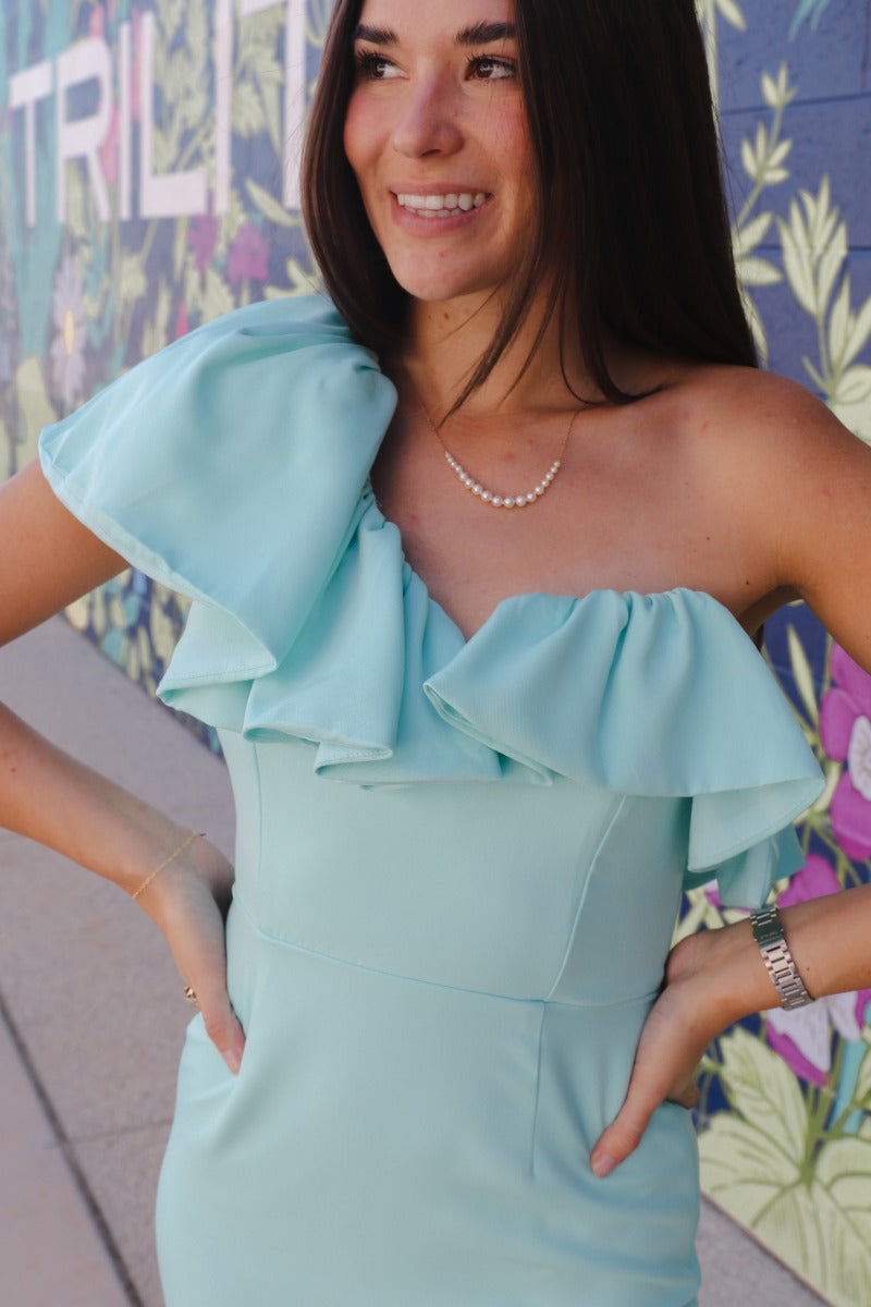 Close up view of model wearing the Level Up Ruffle Dress which features aqua blue fabric, mini length, aqua blue lining, a one shoulder ruffle strap, and a monochromatic side zipper with a hook closure.