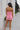 Full body back view of model wearing the I'm Smitten Dress in Pink that has pink ruched mesh fabric, pink lining, a strapless wire v-neck, and a back zipper and hook closure