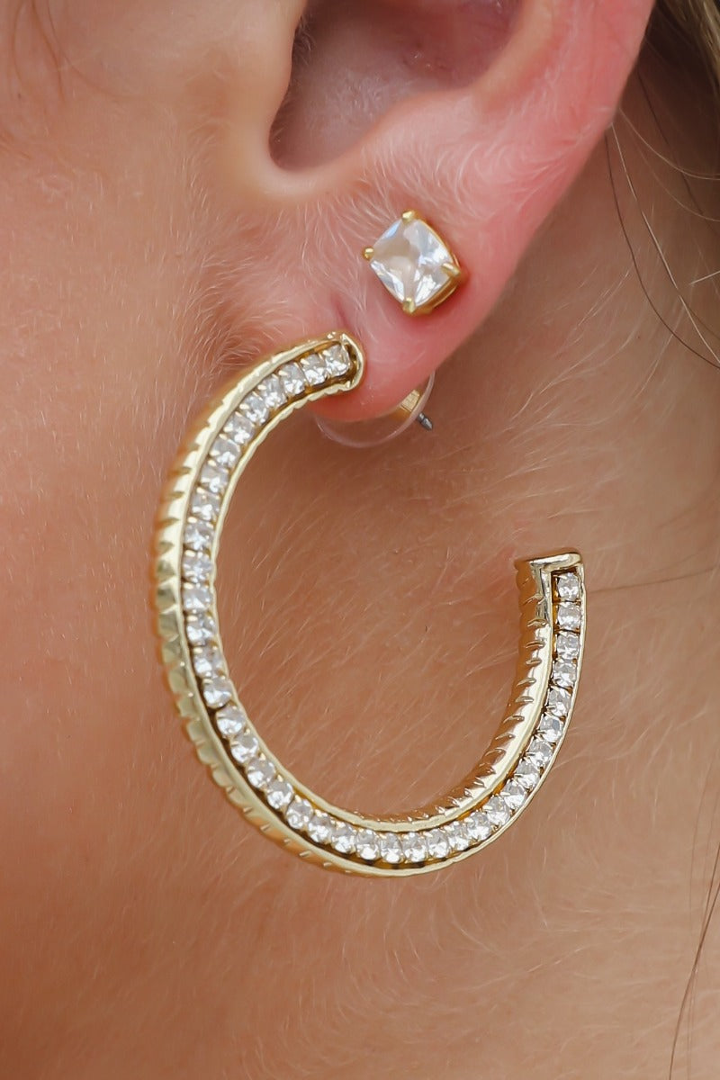 Close up of model wearing the First Class Hoop Earrings that feature medium gold open hoops with clear stones.