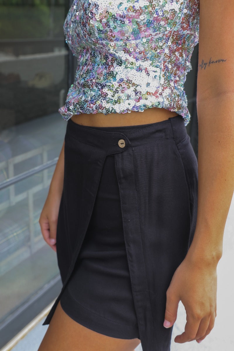 Close up/side view of model wearing the Stepping Out Skort in Black which features black fabric with a slit detail, black shorts lining, a wood button, and a monochromatic side zipper with a hook closure.