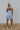Full body back view of model wearing the Now Or Never Dress which features light blue textured fabric, a ruffle two-tiered mini-length body, light blue lining, a square neckline, and thick straps.
