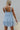 Back view of model wearing the Now Or Never Dress which features light blue textured fabric, a ruffle two-tiered mini-length body, light blue lining, a square neckline, and thick straps.