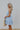 Side view of model wearing the Now Or Never Dress which features light blue textured fabric, a ruffle two-tiered mini-length body, light blue lining, a square neckline, and thick straps.