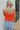 Back view of model wearing the Taste Of The Sun Tank which features red orange fabric, a cowl neckline, red orange lining, and adjustable spaghetti straps.