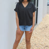 Full body front view of model wearing the For The Record Top which features black fabric, a v neckline, and short sleeves.
