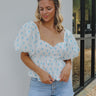 Front view of model wearing the Better Weather Floral Top which features white textured fabric with a light blue floral pattern, a peplum body, a ruched upper with a sweetheart neckline, a smocked back, and short balloon sleeves.