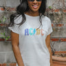 Front view of model wearing the AC/DC Graphic Tee that has white cotton fabric, a round neck, and short sleeves. Graphic says AC/DC in purple, blue, light green, mustard and peach with silver stud embellishments and light blue lining