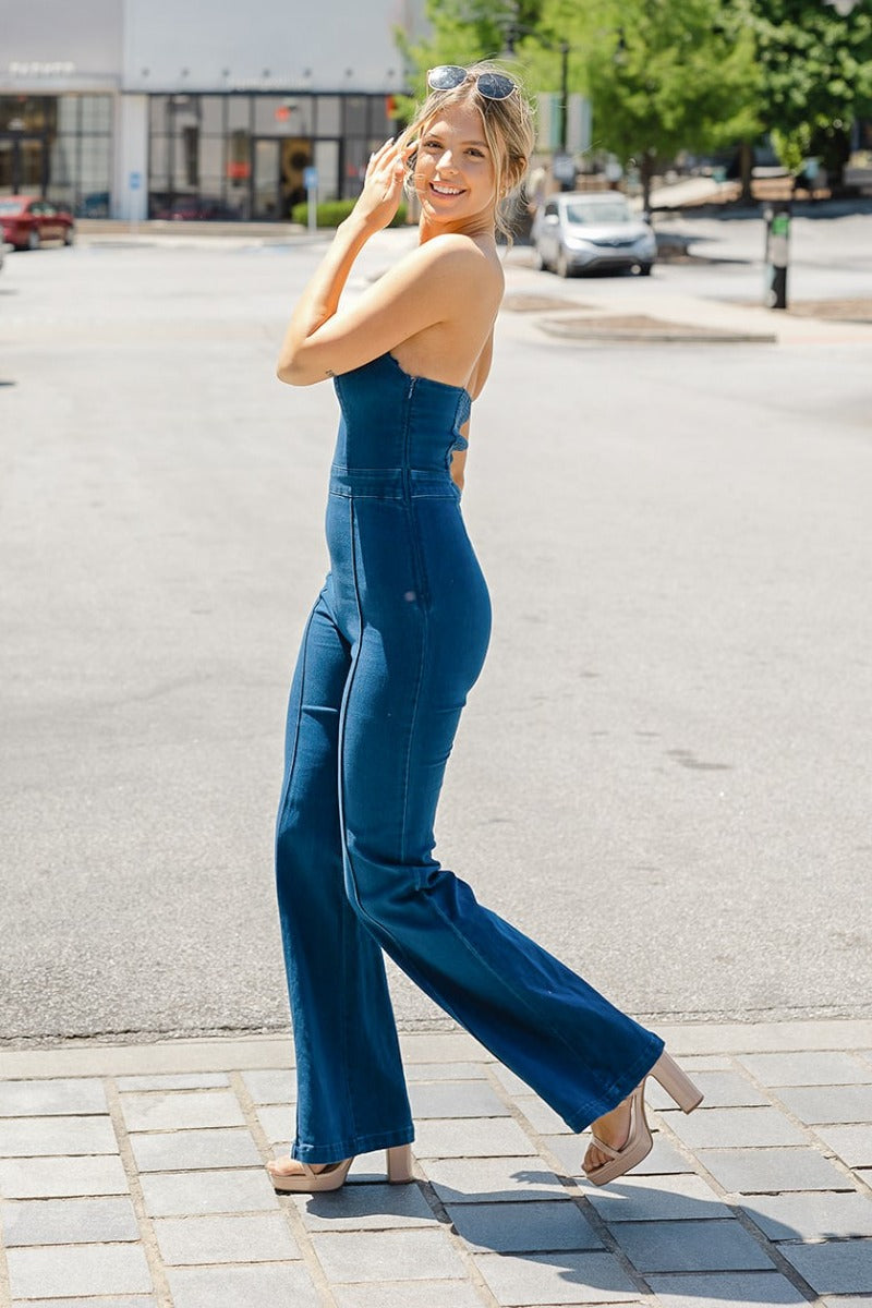 Full side view of model wearing the Daisy Jones Jumpsuit that has dark wash denim fabric, a thick waistband, a strapless sweetheart neck, a smocked back, a side zipper, and flared pant legs