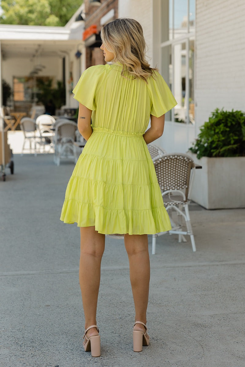 Back view of model wearing the Brighter Days Ahead Dress that has lime green fabric, a three-tiered body style,, a smocked elastic waist, tie details, a v neck with ruffles, and short sleeves
