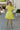 Back view of model wearing the Brighter Days Ahead Dress that has lime green fabric, a three-tiered body style,, a smocked elastic waist, tie details, a v neck with ruffles, and short sleeves