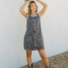 Full body front view of model wearing the Trouble Maker Denim Dress that has charcoal washed denim, front pockets, mini length, gold snap buttons, a round neck, and straps that cross in the back