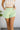 Front view of model wearing the Izzy Denim Shorts in Green that have light green denim fabric, a front zipper and button closure, belt loops, two front pockets, and frayed hem details.