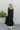 Full body back view of model wearing the Lennon Maxi Dress that has black flowy fabric, a two tiered body style, maxi length, black lining, a square neckline with ruffle trim, and tie straps.