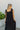 Upper back view of model wearing the Lennon Maxi Dress that has black flowy fabric, a two tiered body style, maxi length, black lining, a square neckline with ruffle trim, and tie straps.