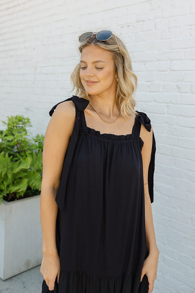 Upper front view of model wearing the Lennon Maxi Dress that has black flowy fabric, a two tiered body style, maxi length, black lining, a square neckline with ruffle trim, and tie straps.