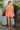 Full body back view of model wearing the Take Me To The Tropics Romper that has coral fabric, an elastic waist, a tie around the waist, an off-the-shoulder neck, and 3/4 flare sleeves with ruffle trim.