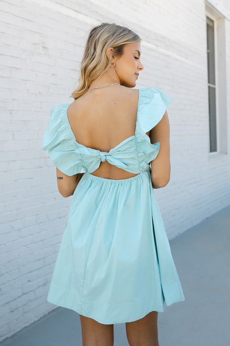 Back view of model wearing the Heart Breaker Dress in Aqua that has aqua fabric with a lining, a mini-length hem, baby doll style, a v neckline, elastic straps with ruffles, and an open back with a bow detail