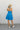 Full body back view of model wearing the Heat Wave Halter Dress that has blue fabric, a two-tiered skirt, mini length, a halter neck with a tie around the neck, an open back with smocking, and a sleeveless design