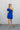 Full body back view of model wearing the In Your Dreams Dress that has royal blue fabric, royal blue lining, a wrap waist, a ruffle hem, one shoulder with a short sleeve, and a monochromatic side zipper with a hook closure.