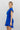 Frontal side view of model wearing the In Your Dreams Dress that has royal blue fabric, royal blue lining, a wrap waist, a ruffle hem, one shoulder with a short sleeve, and a monochromatic side zipper with a hook closure.