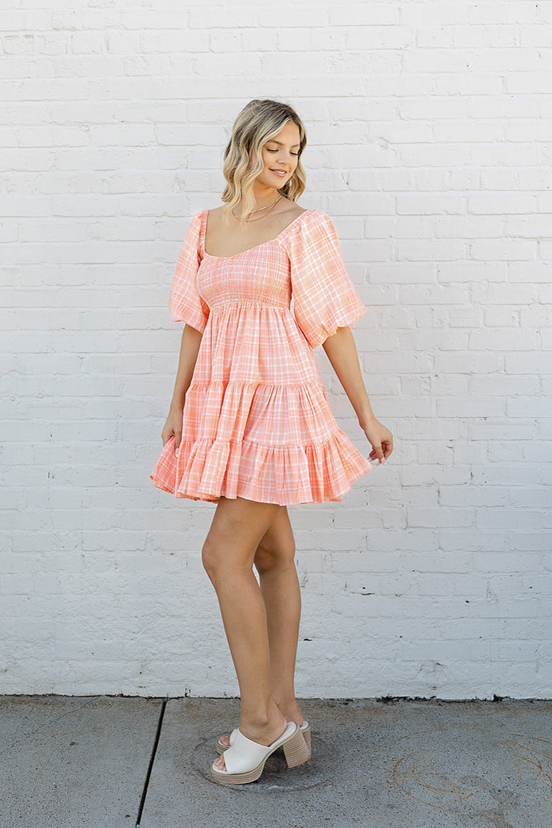 Full body front view of model wearing the Madeline Plaid Dress that has coral fabric with a white plaid pattern, a two-tiered flare skirt, a smocked upper, a sweetheart neck, and short puff sleeves