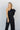 Upper front view of model wearing the Out Of Your League Jumpsuit that has black fabric, black pant lining, an elastic waistband, a one-shoulder neckline with a 3/4 sleeve, and wide pant legs