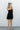 Full body back view of model wearing the On Your Mind Dress that has black fabric with black lining, a one-shoulder neckline, a sleeveless design, a wrap skirt overlay with a side tie, and a back zipper with a hook closure.