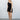 Full body front view of model wearing the On Your Mind Dress that has black fabric with black lining, a one-shoulder neckline, a sleeveless design, a wrap skirt overlay with a side tie, and a back zipper with a hook closure.
