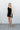 Full body front view of model wearing the On Your Mind Dress that has black fabric with black lining, a one-shoulder neckline, a sleeveless design, a wrap skirt overlay with a side tie, and a back zipper with a hook closure.