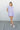 Full body front view of model wearing the Summer In The Air Dress that has lavender gauze fabric, wooden buttons, mini length, pockets on each side, front chest pockets, a collared neckline and short sleeves with cuffs
