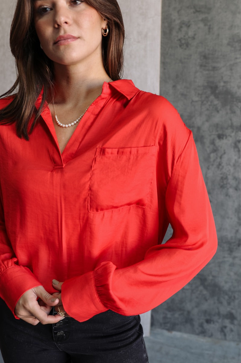 Close-up front view of model wearing the Julianna Red Satin Long Sleeve Top that has red satin fabric, a high-low hem, a chest pocket, a notched collar neck, and long sleeves with buttoned cuffs.