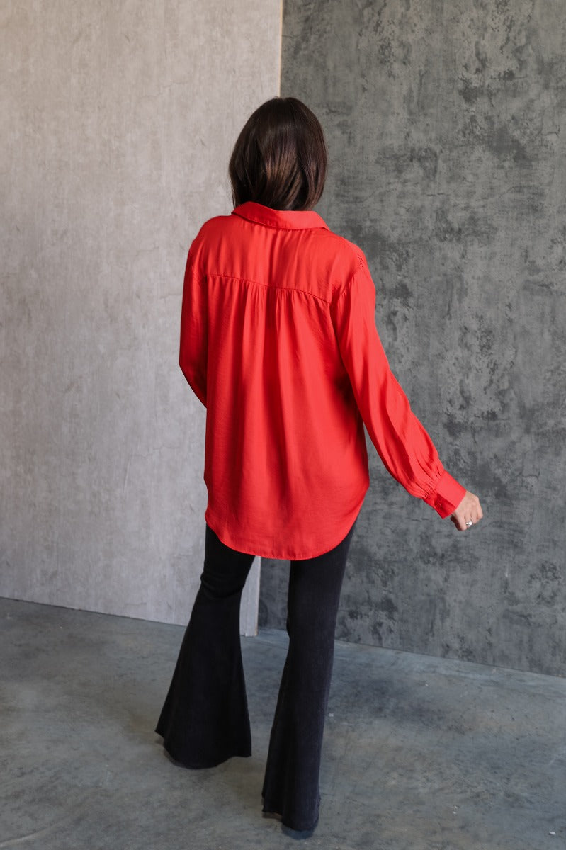 Full body back view of model wearing the Julianna Red Satin Long Sleeve Top that has red satin fabric, a high-low hem, a chest pocket, a notched collar neck, and long sleeves with buttoned cuffs.