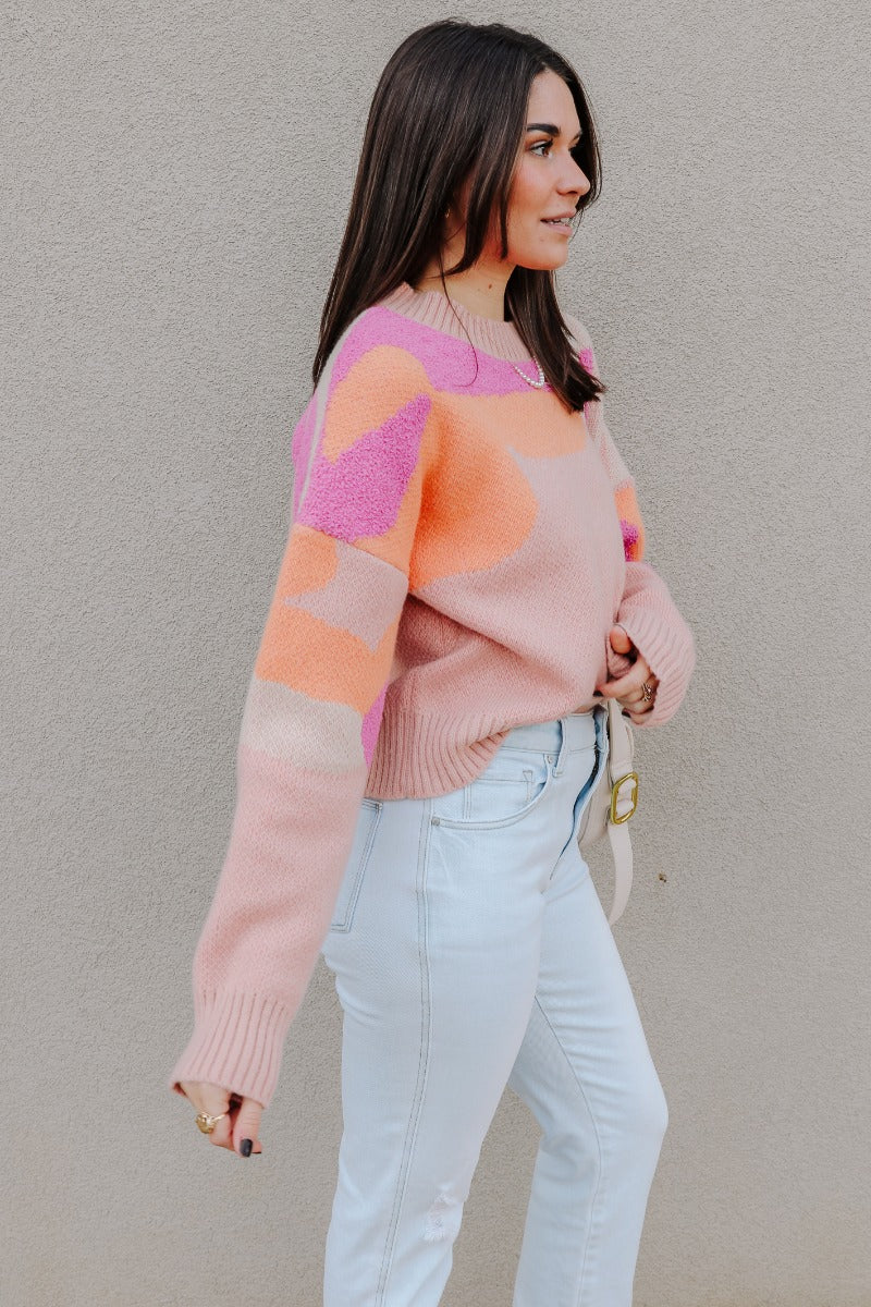 Side view of model wearing the Mila Pink Multi Long Sleeve Sweater which features orange, peach, cream and fuschia knit fabric, an upper floral print, ribbed hem, a ribbed round neckline, dropped shoulders, and long sleeves with ribbed cuffs.