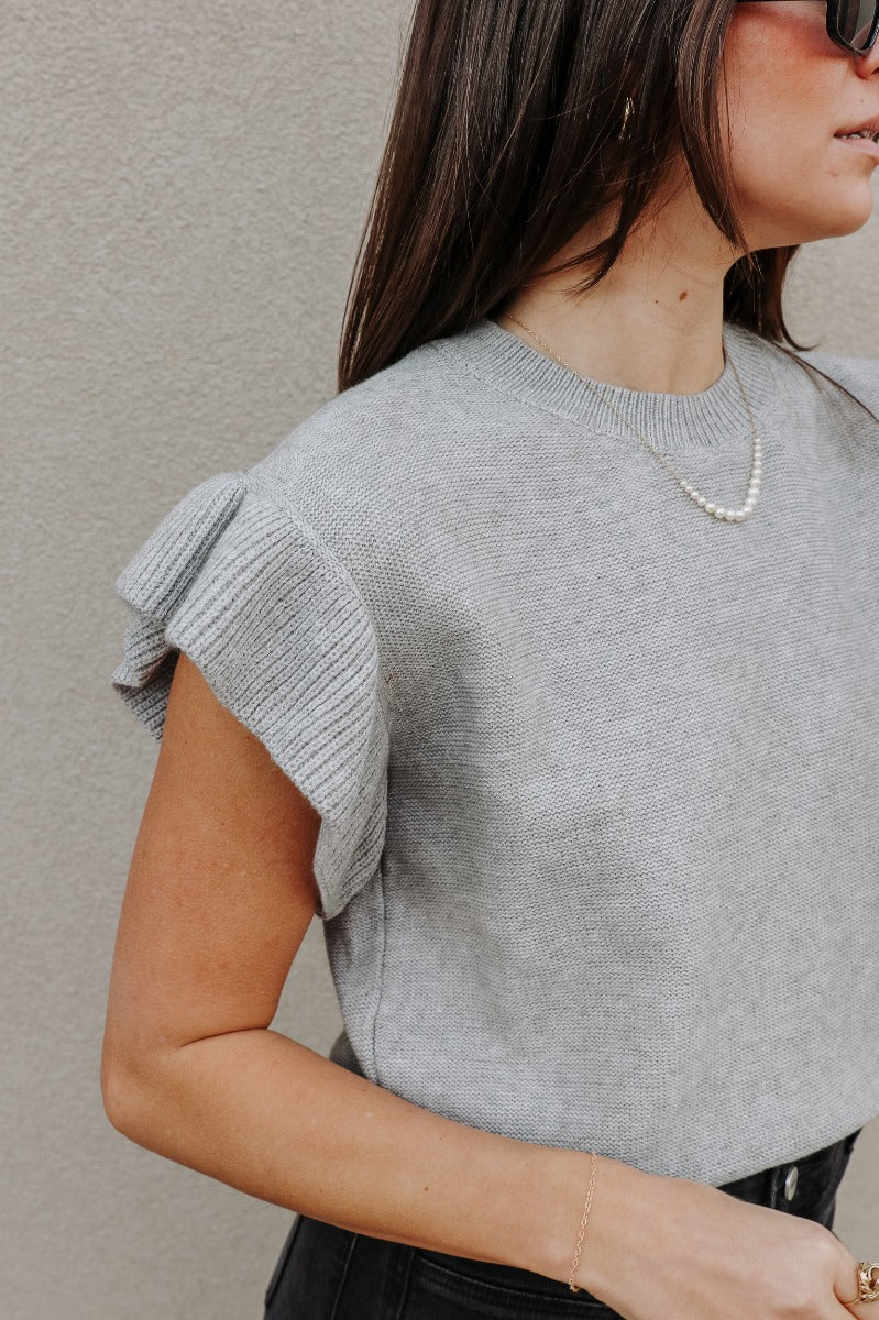 Close up view of model wearing the Sierra Heather Grey Knit Ruffle Sweater which features heather grey knit fabric, ribbed hem, a round neckline, and short sleeves with ruffle details.