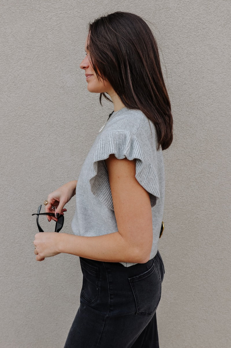 Side view of model wearing the Sierra Heather Grey Knit Ruffle Sweater which features heather grey knit fabric, ribbed hem, a round neckline, and short sleeves with ruffle details.