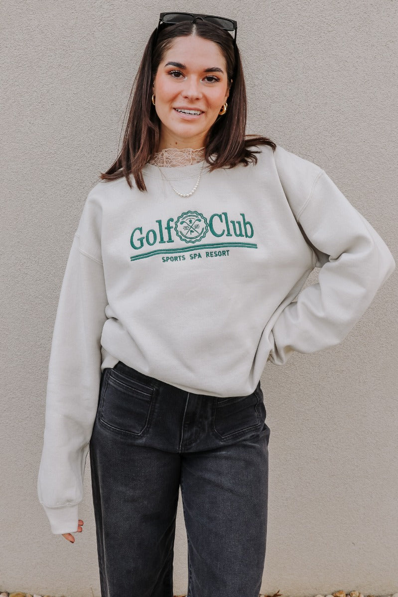 Front view of model wearing the Golf Club Beige & Green Sweatshirt which features beige knit fabric, a round neckline, and long sleeves with ribbed cuffs. Embroidered graphic says "Golf Club" "Sports Spa Resort" with golf clubs symbol in green threading.